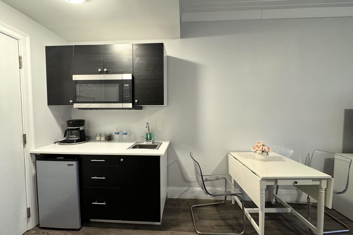 An in-room kitchenette with a microwave set into an overhead cabinet, coffee maker and sink on the counter, an under-counter refrigerator, and a white writing desk with an acrylic chair alongside.