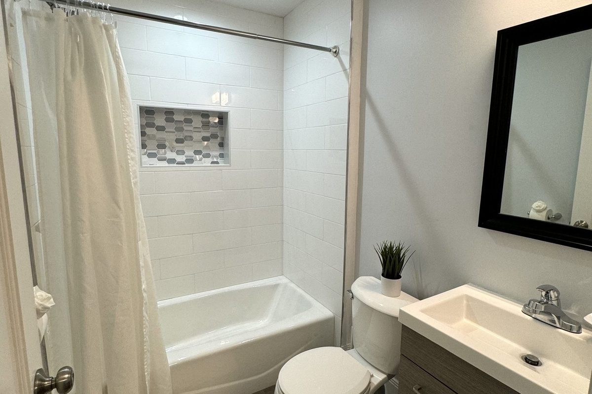 A guest bathroom with a white tub and shower combination, tiled surround with a recessed mosaic shelf, a shower curtain on a curved rod, and a vanity with a black-framed mirror.