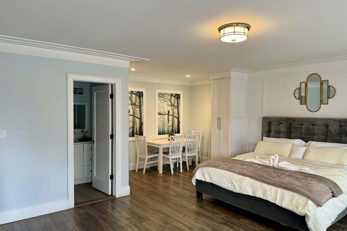 A deluxe king room with a grey upholstered headboard, an open doorway through to the bathroom, and a white dining table with four chairs beside the window.