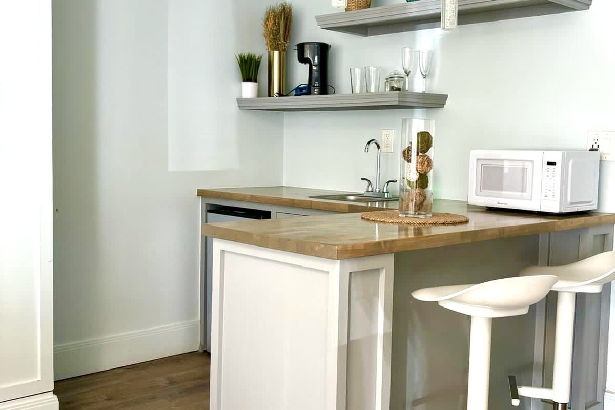 A breakfast bar with a butcher-block counter, two white stools, a microwave and coffee station, and open shelving with plants and glassware above.