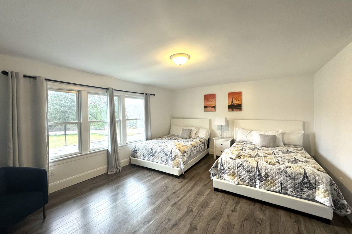Two queen beds with white platform frames and patterned quilts, a nightstand and lamp between them, and wide windows with grey curtains.