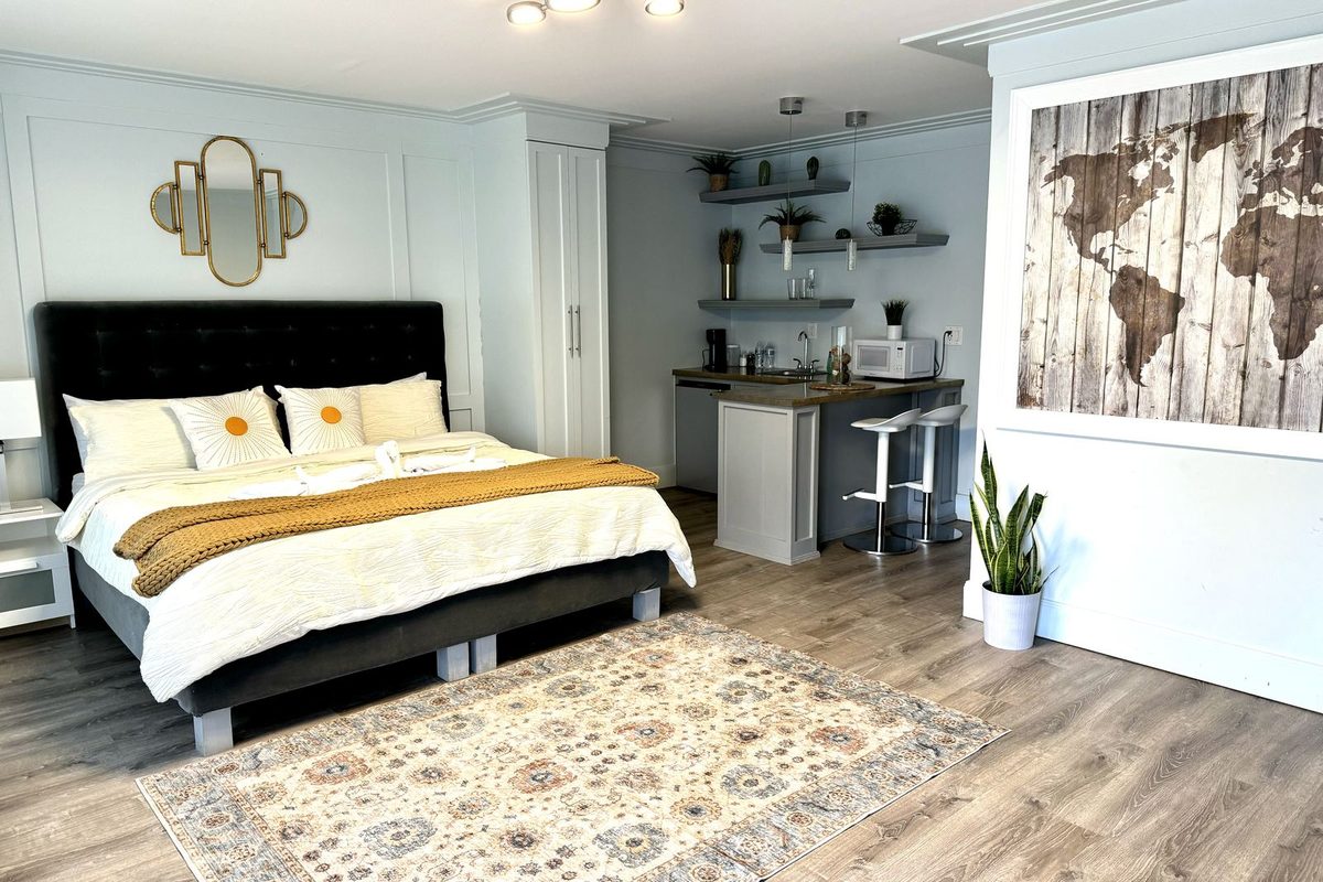 King bed with a tufted charcoal headboard and gold knit throw on a patterned area rug, with a breakfast bar, stools and a microwave in the corner behind the bed.